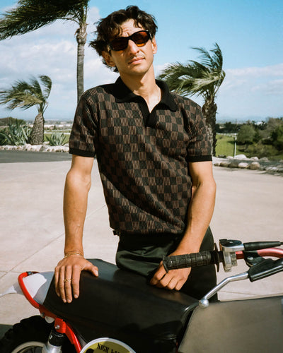 A man in sunglasses and the Dandy Del Mar Sebastian Knit Polo Shirt in Onyx stands outdoors by a dirt bike, with palm trees and a scenic landscape behind him.