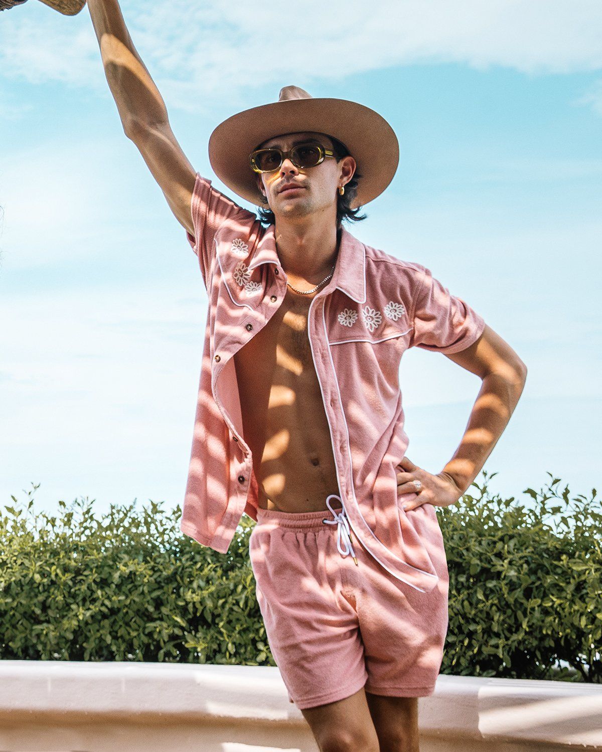 A person in a pink Gaucho Shirt and The Gaucho Shorts in Mauve by Dandy Del Mar, complete with a wide-brim hat and sunglasses, leans against an outdoor wall, surrounded by lush greenery.